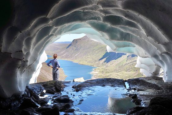 A woman standing at the entance of a snow tunnel on a mountain, looking out towards a river and mountains,