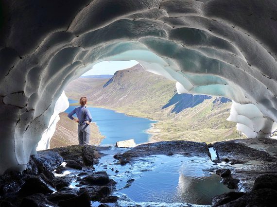 A woman standing at the entance of a snow tunnel on a mountain, looking out towards a river and mountains,