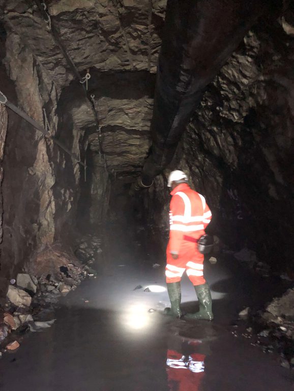 A person dressed in orange high-vis gear and a white helmet seen from behind, walking into a dark, rocky, enclosed space with pipes running along the ceiling.