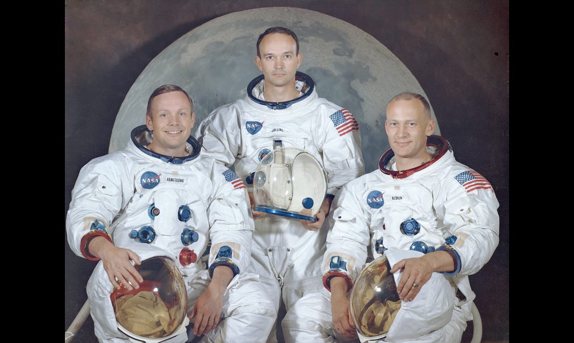 Three male astronauts sit in their astronaut suits with their helmets in their hands in front of an image of the moon.