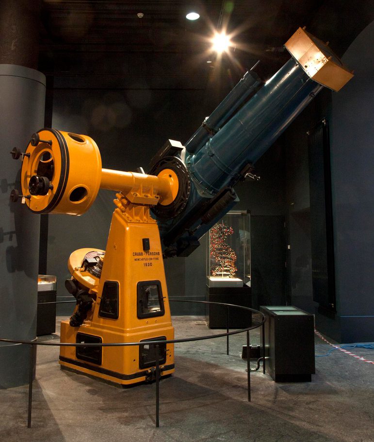 A very large yellow telescope in a building behind barrier ropes.