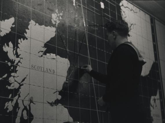 A black and white photograph of a sailor writing on a large map featuring Scotland