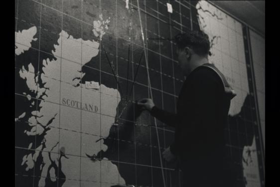 A black and white photograph of a sailor writing on a large map featuring Scotland