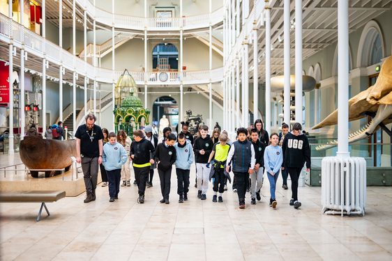 A learning enabler and a big group of school children walk through a large, bright, multi-level gallery space.