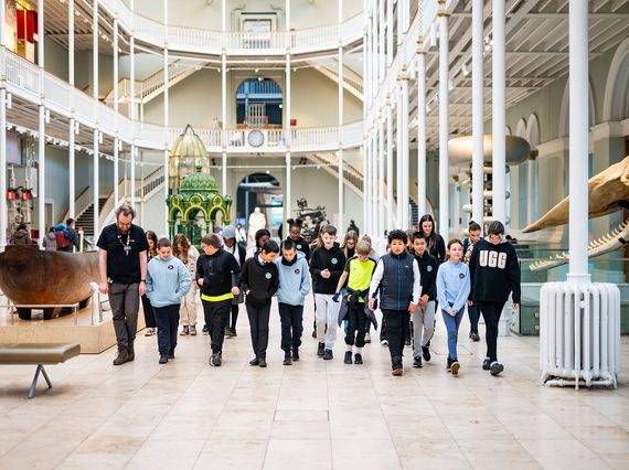 A learning enabler and a big group of school children walk through a large, bright, multi-level gallery space.