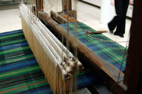 Looking down on a loom mid-weave of a length of blue and green tartan cloth.