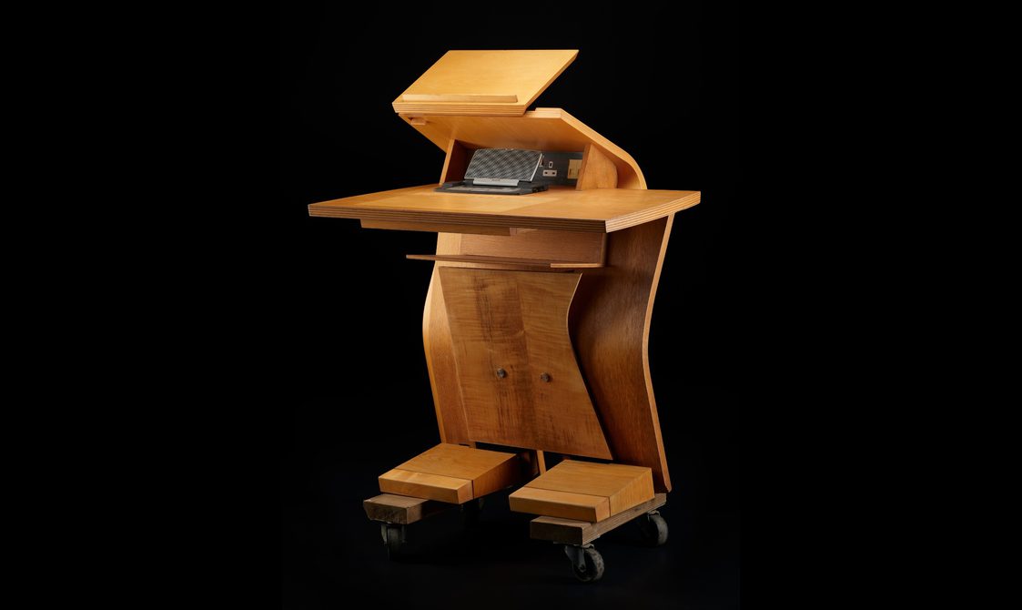 Light brown wooden desk shaped like a humanoid robot, with two blocky 'feet', a tall, curved centre, and two flat surfaces at the top.