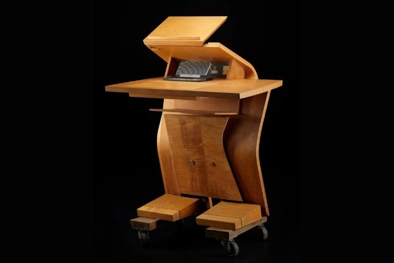 Light brown wooden desk shaped like a humanoid robot, with two blocky 'feet', a tall, curved centre, and two flat surfaces at the top.