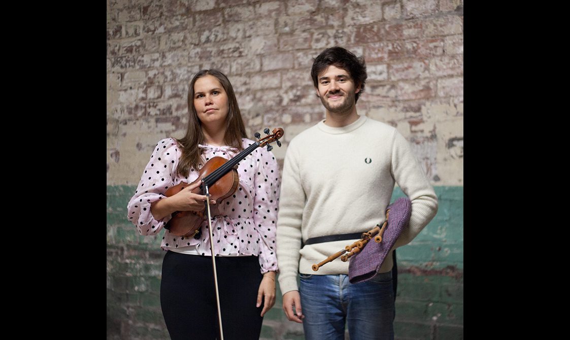A woman holding a fiddle standing beside a man holding the small pipes, both casually dressed and in front of a brick wall..