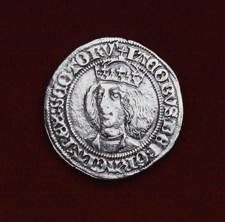 Silver coin, set against red background,with embossed writing to the edge of the coin that centers a bust portrait of a man with shoulder length hair, wearing a crown.