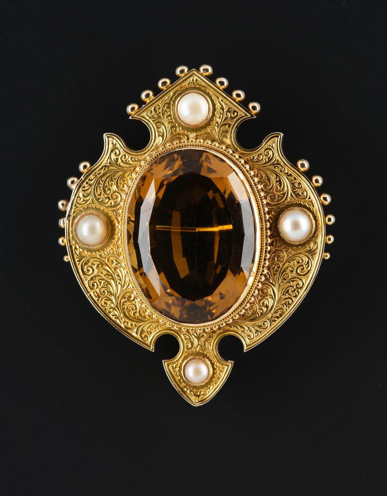 Shield-shaped gold brooch decorated with bead work across the top and sides, with scrolls engraved on the sides and four pearls around a large faceted oval amber stone.