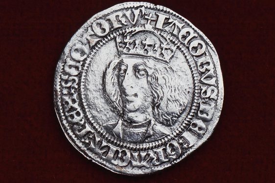 Silver coin, set against red background,with embossed writing to the edge of the coin that centers a bust portrait of a man with shoulder length hair, wearing a crown.
