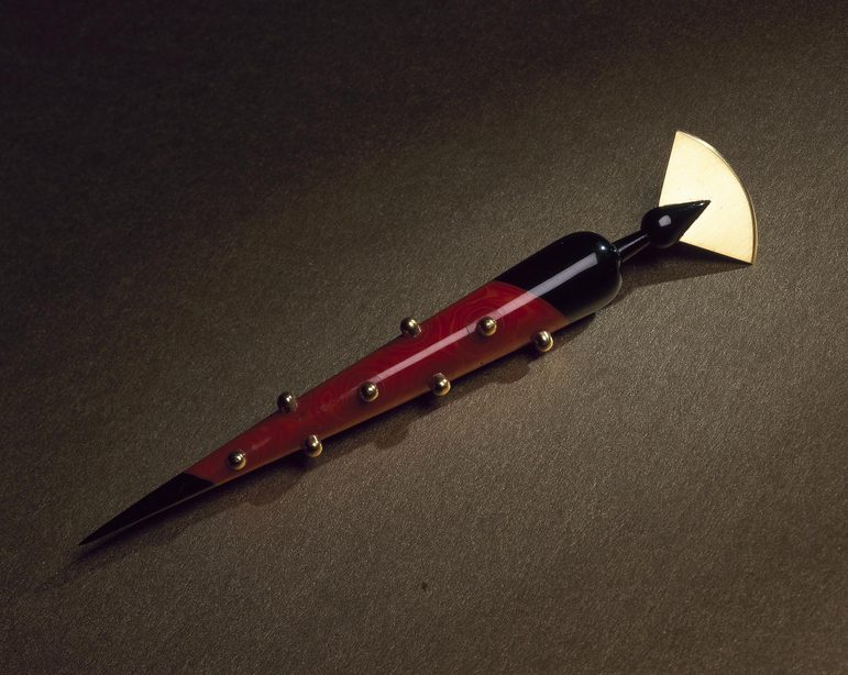 Long conical pen shaped brooch made of red and black plastic covered with gold beading and topped with a gold fan shape.