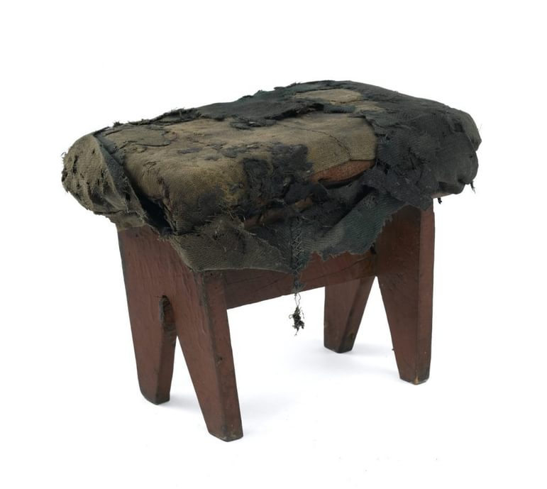 A simple, small and very worn-out wooden stool against a white background. The top is covered by a badly torn and worn green covering.