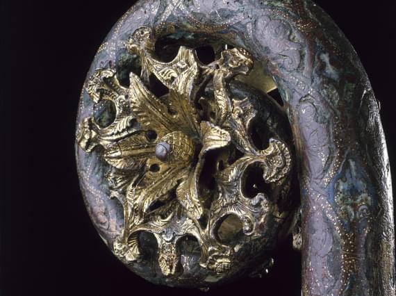 Closeup of metalwork object with a staff and a coiled top. The staff is grey-blue, and a golden sun-like burst is within its coiled top.