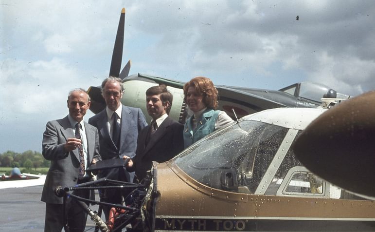 A group of people posing for a photograph next to a Piper PA-24 'Comanche' 260B aircraft. The nose of the aircraft is missing, exposing the inner framework.