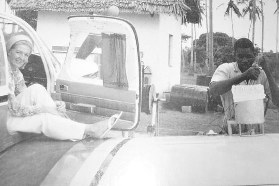 Sheila Scott smiling sitting in the cabin of an aircraft with the door open. Her leg is resting on the wing of the aircraft. A man is filling up the aircraft with fuel.