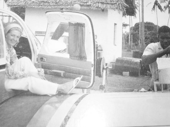 Sheila Scott smiling sitting in the cabin of an aircraft with the door open. Her leg is resting on the wing of the aircraft. A man is filling up the aircraft with fuel.