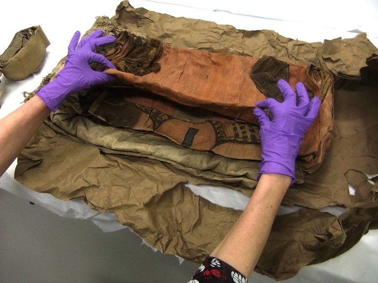 An old looking textile is carefully unwrapped from a brown paper parcel by someone wearing purple gloves.
