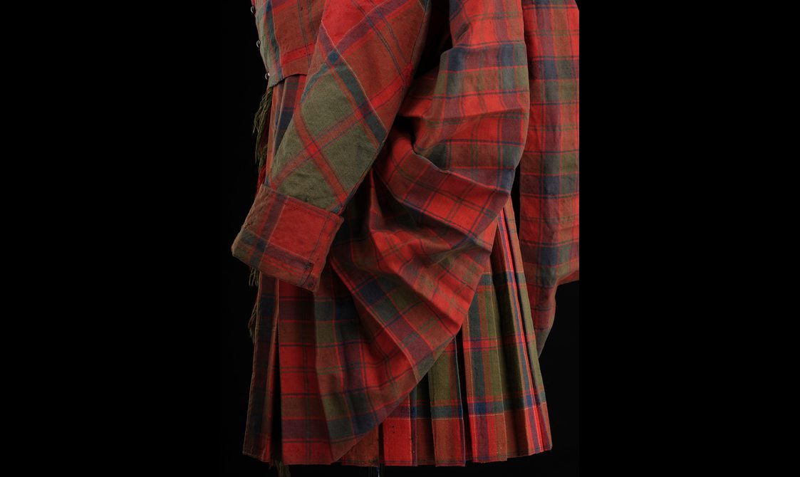 Detail of a mannequin wearing a red pleated tartan kilt with a pinned tail and matching jacket.