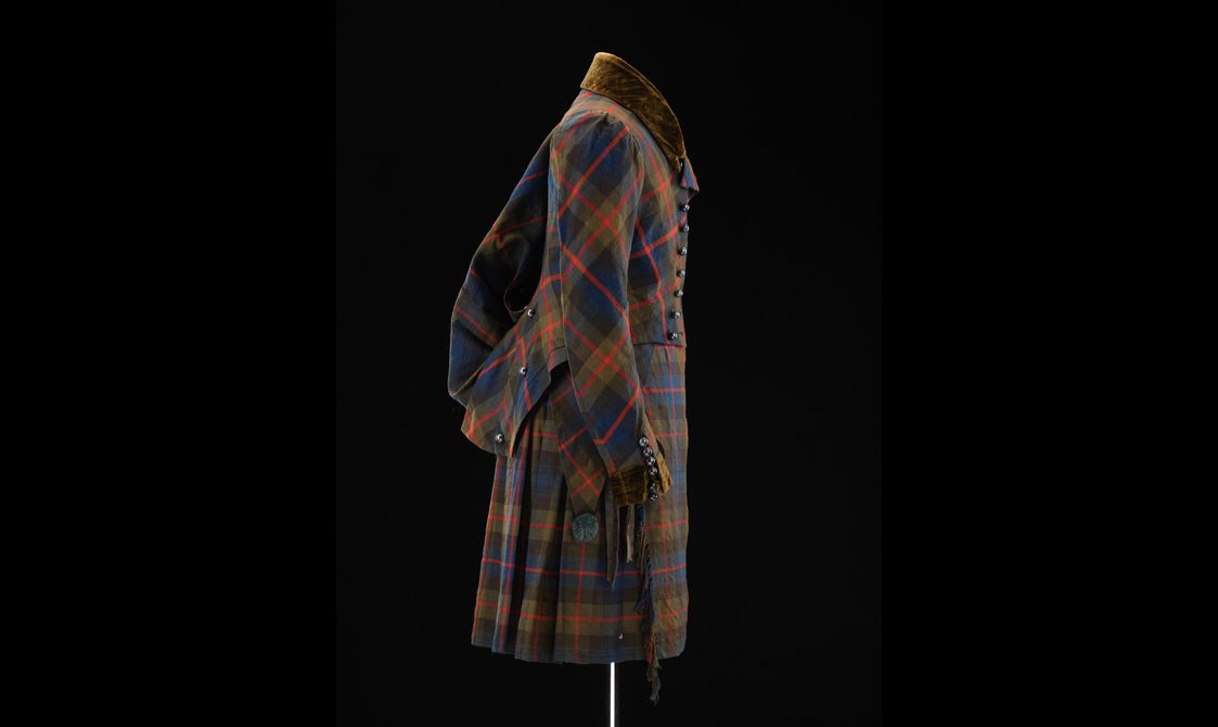 Side view of a mannequin wearing a navy, green, and red tartan kilt and matching jacket with pinned up coat tails.