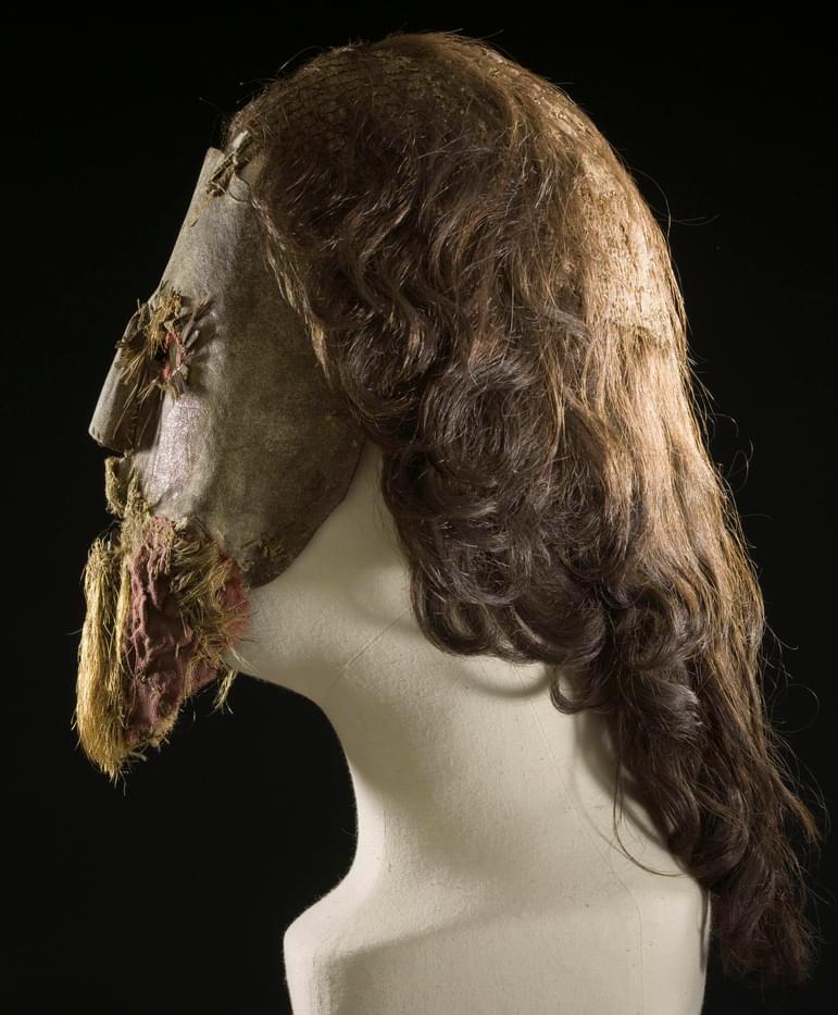 A side view of a mannequin head wearing a full leather face mask with long hair and a beard attached.