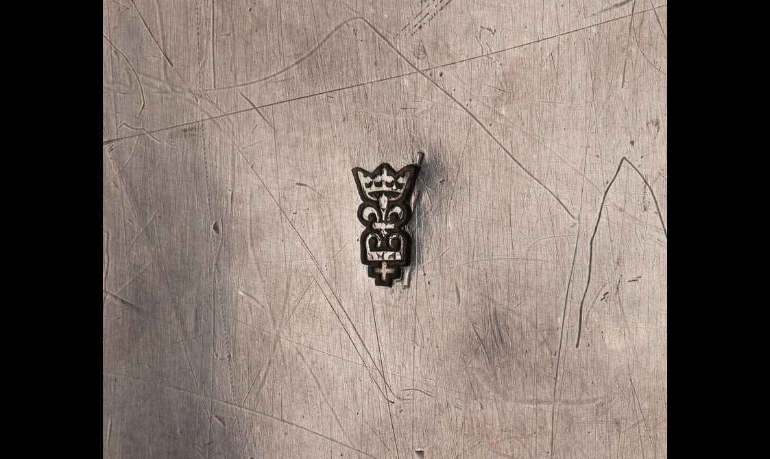 Extreme closeup of a maker's mark on a silver surface. A crown atop a fleur-de-lis atop another crown and a cross.