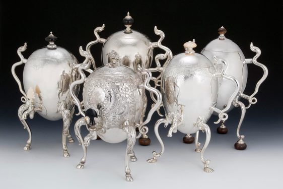 Five large silver coffee pots shaped like eggs propped up on tall, curving legs and with spindly arms, some of them made to resemble serpents. They are very shiny.
