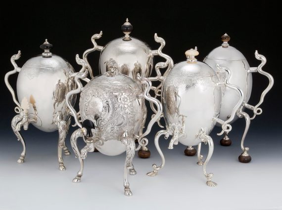 Five large silver coffee pots shaped like eggs propped up on tall, curving legs and with spindly arms, some of them made to resemble serpents. They are very shiny.