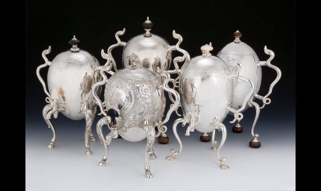 Five large silver coffee pots shaped like eggs propped up on tall, curving legs and with spindly arms, some of them made to resemble serpents. They are very shiny.