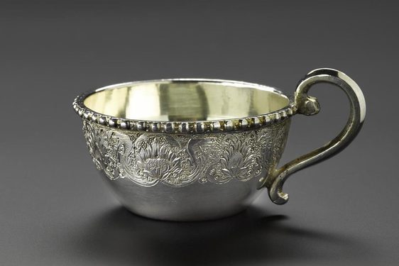 A silver teacup with floral patterns engraved on its surface with a curved handle.