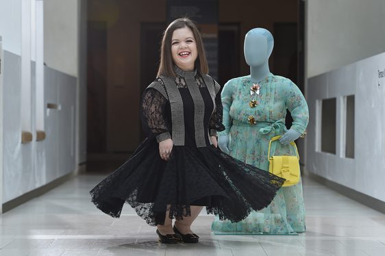 A smiling woman in a black dress with a flared skirt standing beside a mannequin wearing a green floral dress.
