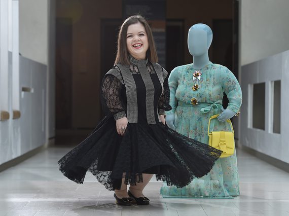 A smiling woman in a black dress with a flared skirt standing beside a mannequin wearing a green floral dress.