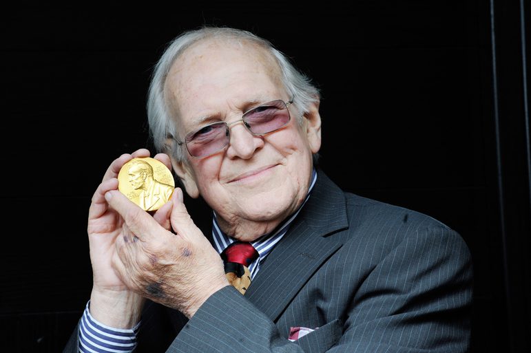 A man with white hair and tinted glasses wearing a suit jacket, blue striped shirt, and red tie holds a gold nobel prize medal in both hands close to this face.
