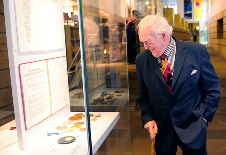A man with white hair and glasses wearing a navy suit and colourful tie looks into a museum display case