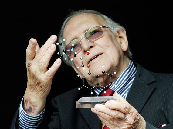 A man with white hair and glasses wearing a suit holds a model of a molecular structure