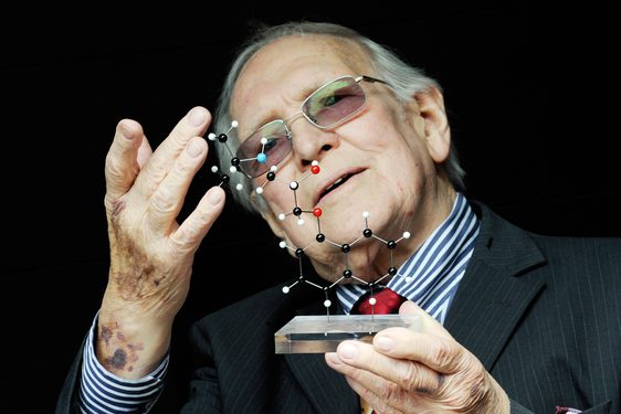 A man with white hair and glasses wearing a suit holds a model of a molecular structure