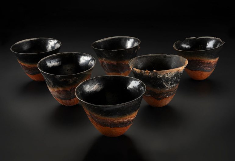 A set of six small black and terracotta cups. They are irregular in shape and sit against a black background