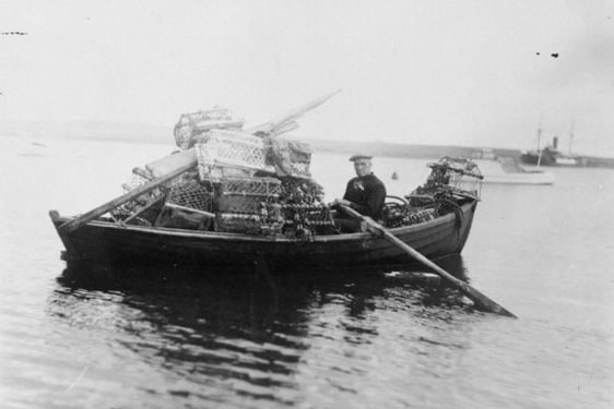 Person sitting in a wooden boat filled with lobster traps on calm water, with another boat visible in the background.