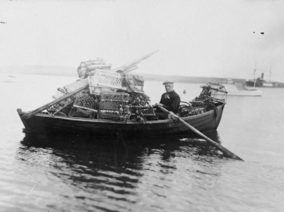 Person sitting in a wooden boat filled with lobster traps on calm water, with another boat visible in the background.
