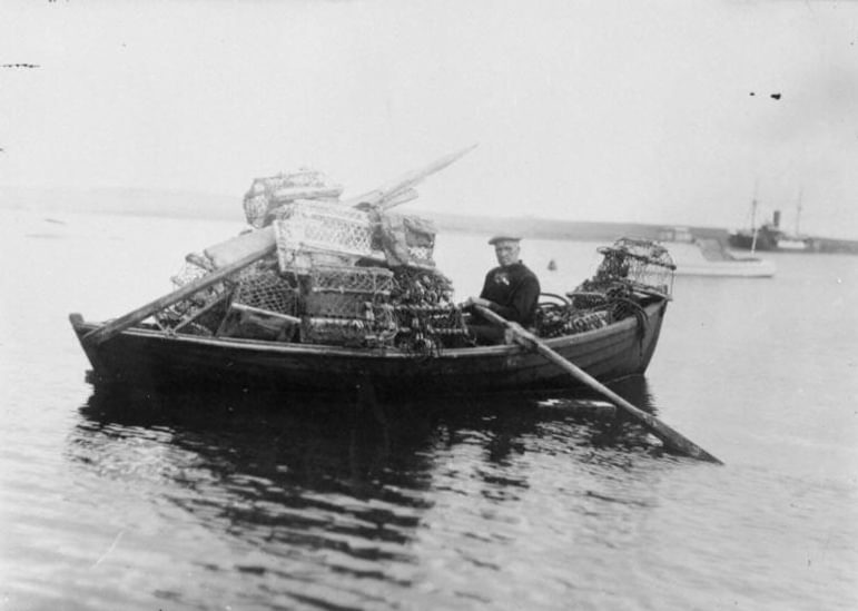 Person sitting in a wooden boat filled with lobster traps on calm water, with another boat visible in the background.