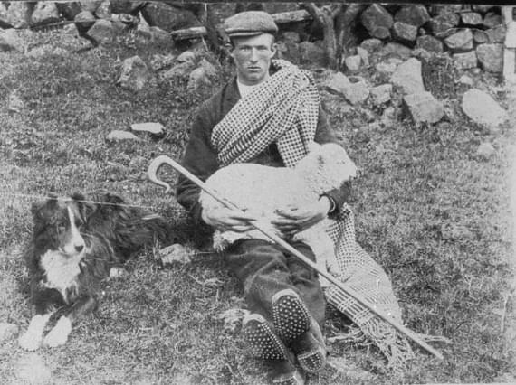 A young shepherd sits on the ground near a stone wall. He is holding a lamb on his knees. A sheepdog sits near him.