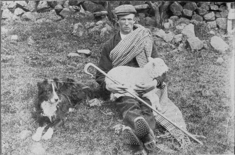 A young shepherd sits on the ground near a stone wall. He is holding a lamb on his knees. A sheepdog sits near him.