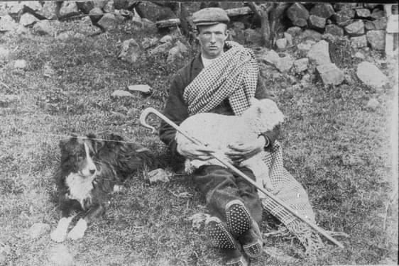 A young shepherd sits on the ground near a stone wall. He is holding a lamb on his knees. A sheepdog sits near him.
