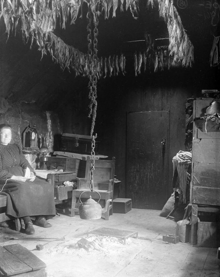 Old black and white image of a woman sitting by a central fire with a kettle hanging over it inside a traditional, rustic stone and timber house.