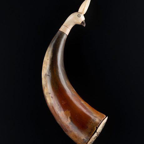 A container made from a cow’s brown horn fitted with an ivory carving of a bird’s head. The container is photographed against a black background.