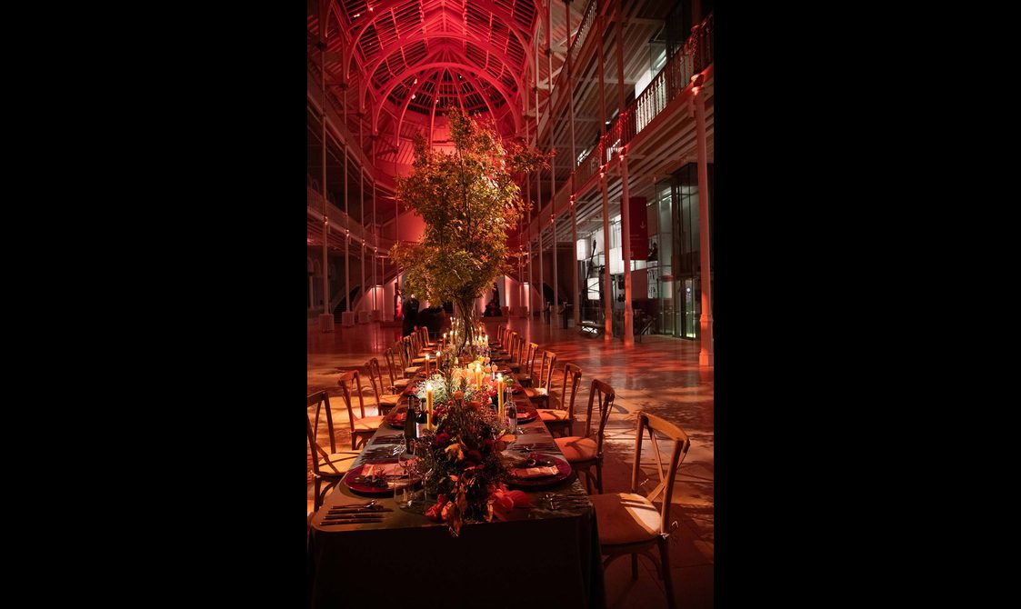 A long table set up for dinner in a multi-level museum gallery with a domed ceiling. There is a tree at the end of the table and the space is lit with red lights.