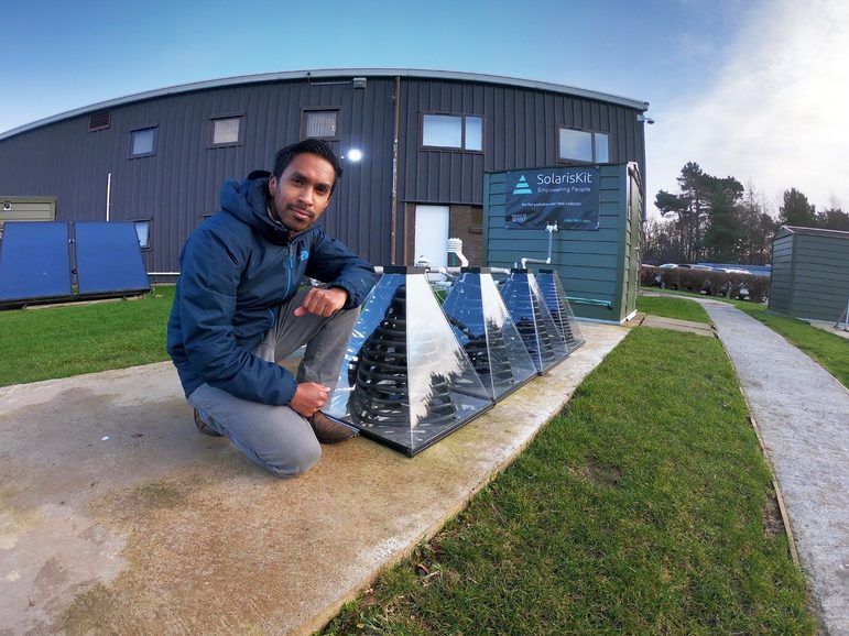 A man in a blue hooded jacket crouches in front of a row of transparent pyramid shaped objects with coils inside. They are outside a building that has a 'SolarisKit' sign on it.