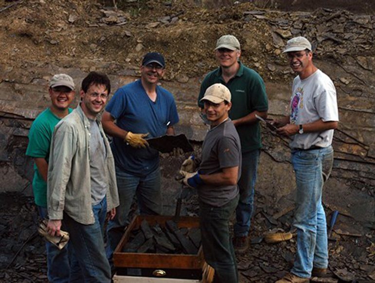 Six men in a rocky quarry, five of them wearing baseball hats, turn to smile at the camera.