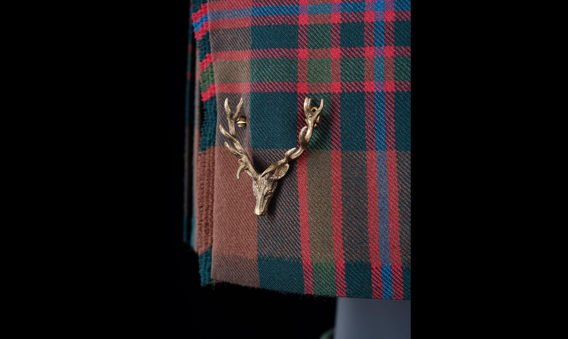 A detail of a red, green, and blue tartan kilt with a pin of a stag head.